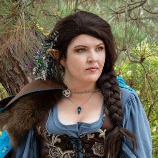 Ren is standing in front of a blurry bookshelf. They have a smoky eye with a gold corner, are wearing black lipstick, and their brown hair falls in loose curls at their chin. Half of their face is obstructed by their right hand holding d20s between their f